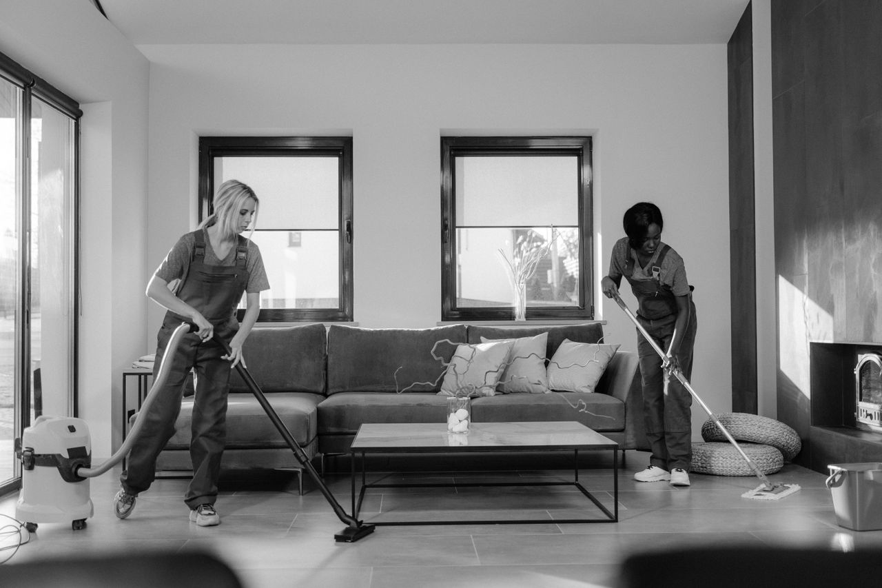 Two women dressed for work are cleaning a contemporary living room with a vacuum cleaner and a mop.