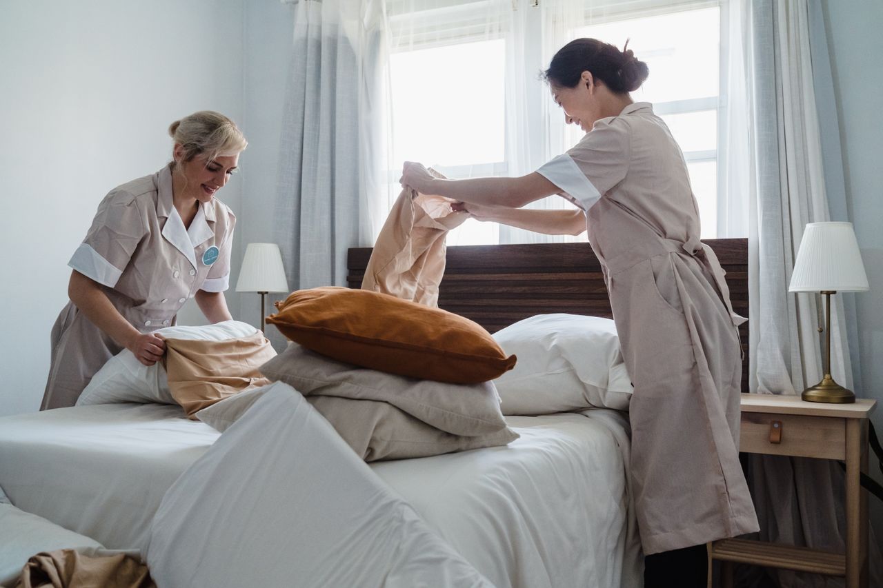 Two housekeepers in uniform fixing a hotel room bed with fresh linen and pillows.